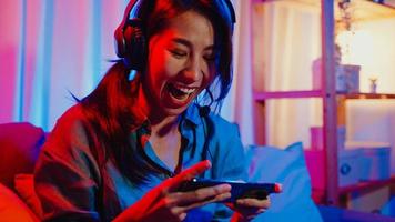 videojuego de competencia de auriculares de uso de jugador de niña de Asia feliz en línea con teléfono inteligente charla emocionada con un amigo sentado en el sofá en la sala de estar de luces de neón de colores en casa, concepto de actividad de cuarentena en casa. foto