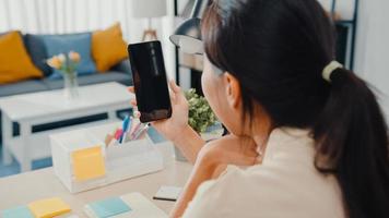 La jovencita asiática usa un teléfono inteligente con pantalla negra en blanco simulada para mostrar texto publicitario mientras trabaja de manera inteligente desde casa en la sala de estar. tecnología chroma key, concepto de diseño de marketing. foto
