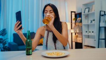joven asiática bebiendo cerveza divirtiéndose feliz momento fiesta nocturna evento de año nuevo celebración en línea a través de videollamada por teléfono en casa por la noche. distanciamiento social, cuarentena para la prevención del coronavirus.