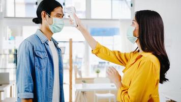 Asia female receptionist conducting wear protective face mask use infrared thermometer checker or temperature gun on customer's forehead before enter office. Lifestyle new normal after corona virus. photo