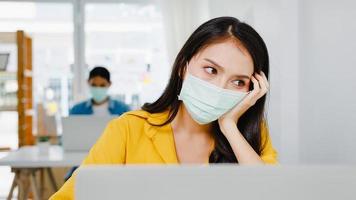 Freelance Asia women wear face mask using laptop hard work at new normal home office. Working from house overload, self isolation, social distancing, quarantine for corona virus prevention. photo