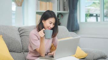 Mujer asiática independiente de negocios joven que trabaja en la computadora portátil revisando las redes sociales y tomando café mientras está acostado en el sofá cuando se relaja en la sala de estar en casa. mujeres de estilo de vida en el concepto de casa.