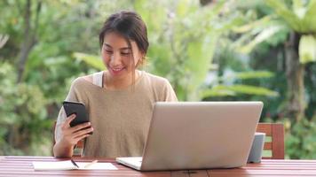 Freelance Asian woman working at home, business female working on laptop and using mobile phone drinking coffee sitting on table in the garden in morning. Lifestyle women working at home concept.