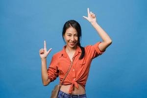 Portrait of young Asian lady smiling with cheerful expression, shows something amazing at blank space in casual cloth and looking at camera isolated over blue background. Facial expression concept. photo