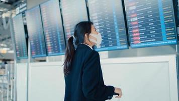 Asian business girl wear face mask with suitcase stand in front of board look at information checking her flight at international airport. Business commuter covid pandemic, Business travel concept. photo