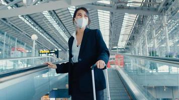 Asian business girl wear face mask drag luggage stand on escalator look around walk to terminal at international airport. Business commuter covid pandemic, Business travel social distancing concept. photo