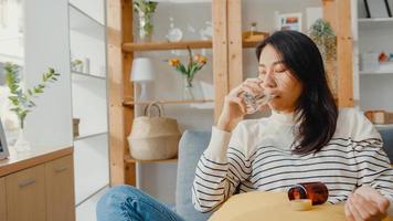 mujer joven enferma de asia sosteniendo un vaso de agua con pastillas tomar la medicina sentarse en el sofá en casa. chica tomando medicamentos después de la orden del médico, cuarentena en la casa, concepto de cuarentena de distanciamiento social de coronavirus. foto