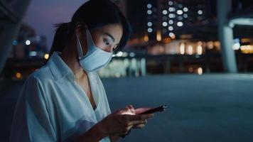 Joven empresaria asiática en ropa de moda con mascarilla usando un teléfono inteligente escribiendo un mensaje de texto mientras está parado al aire libre en la ciudad urbana por la noche. distanciamiento social para evitar la propagación del concepto covid-19. foto