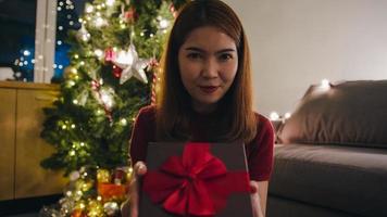 Young Asia female using mobile phone video call talking with couple with X'Mas present box, Christmas tree decorated with ornament in living room at home. Christmas night and New Year holiday festival