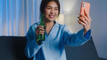 joven asiática bebiendo cerveza divirtiéndose feliz momento fiesta nocturna evento celebración en línea a través de videollamada en la sala de estar en casa por la noche. distanciamiento social, cuarentena para la prevención del coronavirus.