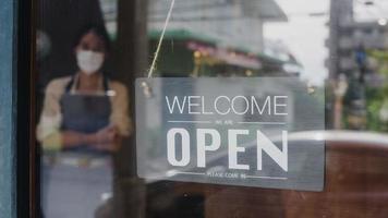 Young Asia girl wear face mask turning a sign from closed to open sign on door looking outside waiting for clients after lockdown. Owner small business, food and drink, business reopen again concept. photo