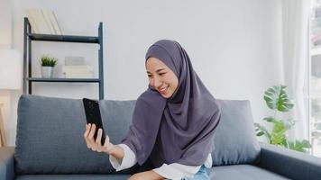 Asia dama musulmana usa hijab usando videollamada telefónica hablando con pareja en casa. joven adolescente haciendo video de vlog en las redes sociales en el sofá de la sala de estar. distanciamiento social, cuarentena por coronavirus. foto