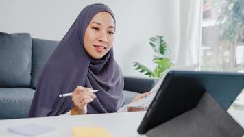 Asia muslim lady wear headscarf casual use tablet talk to colleagues about sale report in conference video call while working from home at living room. Social distancing, quarantine for corona virus. photo