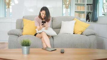 Young Asian woman using smartphone checking social media feeling happy smiling while lying on the sofa when relax in living room at home. Lifestyle latin and hispanic ethnicity women at house concept. photo