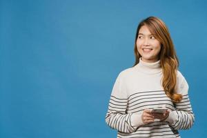 Young Asia lady using phone with positive expression, smiles broadly, dressed in casual clothing feeling happiness and standing isolated on blue background. Happy adorable glad woman rejoices success. photo