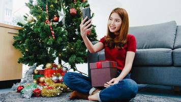 Young Asian female using smart phone video call talking with couple with X'Mas present box, Christmas tree decorated with ornament in living room at home. Christmas and New Year holiday festival. photo