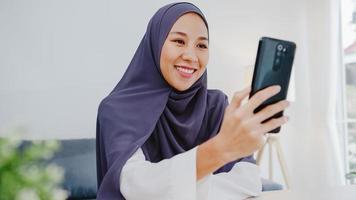 Joven empresaria musulmana asiática que usa un teléfono inteligente, habla con un amigo por videochat y realiza una lluvia de ideas en línea mientras trabaja de forma remota desde su casa en la sala de estar. distanciamiento social, cuarentena por coronavirus. foto