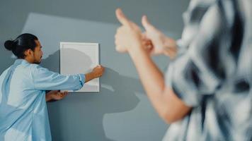 Feliz pareja joven y atractiva asiática hombre y mujer se ayudan mutuamente a colgar el marco de la imagen en la pared para decorar la casa y el paquete de caja de cartón en la sala de estar. concepto de hogar de decoración asiática joven casada. foto