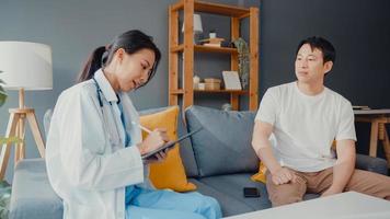 Young Asia female professional physician doctor using digital tablet sharing good health test news with happy male patient sit on couch in house. Medical insurance, Visit patient at home concept. photo