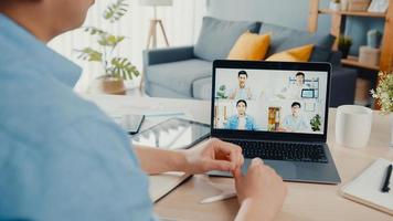 Young Asia businessman using laptop talk to colleagues about plan in video call meeting while work from home at living room. Self-isolation, social distancing, quarantine for corona virus prevention.