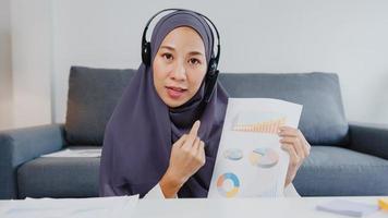 Asia muslim lady wear headphone using computer laptop talk to colleagues about sale report in video call while remotely work from home at living room. Social distancing, quarantine for corona virus. photo