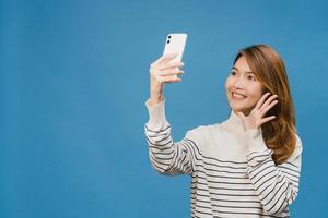 Smiling adorable Asian female making selfie photo on smart phone with positive expression in casual clothing and stand isolated on blue background. Happy adorable glad woman rejoices success.