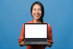 Young Asia lady show empty laptop screen with positive expression, smiles broadly, dressed in casual clothing feeling happiness isolated on blue background. Computer with white screen in female hand. photo