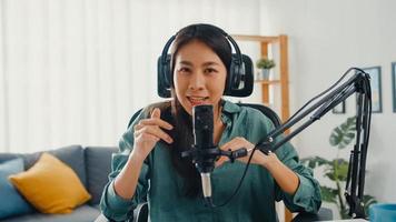 feliz niña asiática grabar un podcast con auriculares y micrófono, mirar a la cámara, hablar y descansar en su habitación. Podcaster femenina hace podcast de audio desde su estudio en casa, permanece en el concepto de casa. foto
