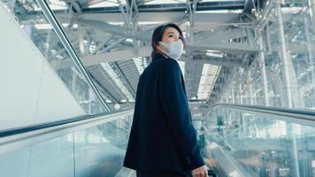 Asian business girl wear face mask drag luggage stand on escalator look around walk to terminal at international airport. Business commuter covid pandemic, Business travel social distancing concept. photo