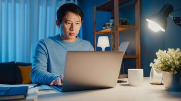 Asia empresario independiente se concentra en trabajar escribiendo en una computadora portátil en línea de forma remota desde la empresa en el escritorio en la sala de estar en casa horas extras por la noche, trabajar desde casa durante el concepto de pandemia covid-19.