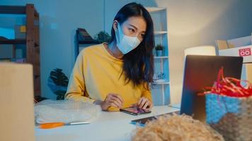Young Asia businesswoman wear face mask check purchase order on smartphone and stock in computer in home office at night. Small business owner, online market delivery, lifestyle freelance concept.