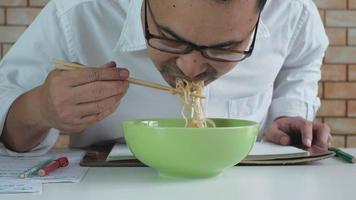 trabajador de sexo masculino asiático lee un libro de citas mientras come fideos instantáneos en un tazón verde con palillos en la mesa en la oficina de fondo de pared de ladrillo durante un descanso a la hora del almuerzo, un estilo de vida apresuradamente insalubre. video