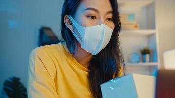 Young Asia businesswoman wear face mask check purchase order and reply client inbox chat on laptop in home office at night. Small business owner, online market delivery, lifestyle freelance concept.