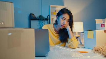 Young Asia businesswoman look around room full of product stuff and parcel box feel stress and upset with bad sell in home office at night. Small business owner, online market delivery concept. photo