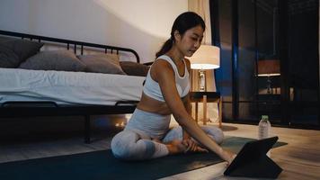 joven asiática en ejercicios de ropa deportiva haciendo ejercicio y usando la tableta para ver el video tutorial de yoga en la noche en casa. entrenamiento a distancia con entrenador personal, distancia social, concepto de educación en línea.