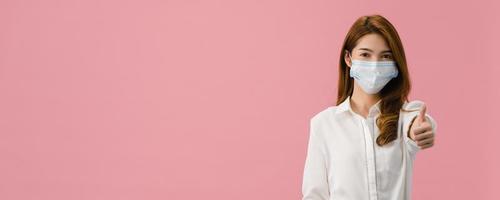 Young Asia girl wearing face mask showing thumb up with dressed in casual cloth and look at camera. Social distancing, quarantine for corona virus. Panoramic banner pink background with copy space. photo