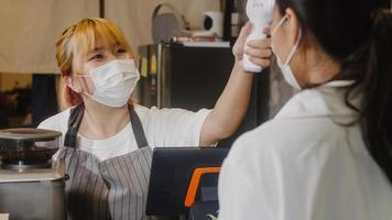 Young Asia female restaurant staff wearing protective face mask using infrared thermometer checker or temperature gun on customer's forehead before enter. Lifestyle new normal after corona virus.