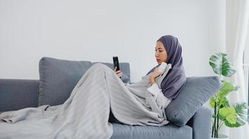 joven musulmana de asia usa hijab usando videollamada telefónica hablando con consulta médica o consulta en línea en el sofá de la sala de estar en casa. distanciamiento social, cuarentena por concepto de coronavirus. foto