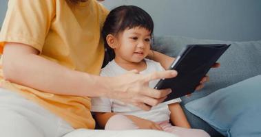 Happy cheerful Asia family mom and cute kid using digital tablet interest cartoon and watch movie having fun relax on couch in living room at house. Spending time together, Quarantine for coronavirus. photo