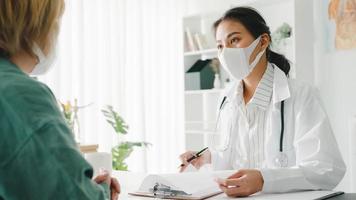 Young Asia lady doctor wear protective mask using clipboard is delivering great news talk discuss results or symptoms with girl patient in hospital office. Lifestyle new normal after corona virus. photo