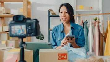 Young Asia lady fashion designer using mobile phone receiving purchase order and show clothes recording video live streaming online with camera. Small business owner, online market delivery concept. photo