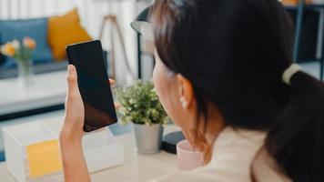 La jovencita asiática usa un teléfono inteligente con pantalla negra en blanco simulada para mostrar texto publicitario mientras trabaja de manera inteligente desde casa en la sala de estar. tecnología chroma key, concepto de diseño de marketing. foto