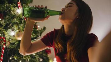 Young Asia female drinking beer having fun happy night party video call talk with couple, Christmas tree decorated with ornaments in living room at home. Christmas night and New Year holiday festival. photo