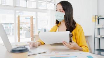 Asia businesswoman entrepreneur wearing medical face mask for social distancing in new normal situation for virus prevention while using laptop back at work in office. Lifestyle after corona virus. photo