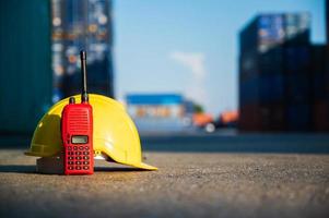 Radio walkie talkie and helmet hard hat on the floor at Container cargo harbor with sunlight background. Business Logistics import export shipping concept.