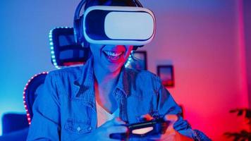 Happy young asia girl gamer winner wear virtual reality glasses goggles headset and joystick controller fun and excited with online game in neon home studio at night, Home quarantine activity. photo