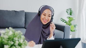 Asia mujer musulmana usa auriculares con tableta hable con sus colegas sobre el informe de venta en una videoconferencia mientras trabaja desde su casa en la sala de estar. distanciamiento social, cuarentena por coronavirus.