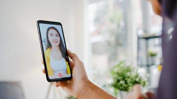 Joven empresaria musulmana asiática que usa un teléfono inteligente, habla con un amigo por videochat y realiza una lluvia de ideas en línea mientras trabaja de forma remota desde su casa en la sala de estar. distanciamiento social, cuarentena por coronavirus. foto