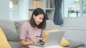 Asian woman using laptop and credit card shopping ecommerce, female relax feeling happy online shopping sitting on sofa in living room at home. Lifestyle women relax at home concept.