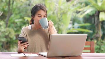 Freelance Asian woman working at home, business female working on laptop and using mobile phone drinking coffee sitting on table in the garden in morning. Lifestyle women working at home concept. photo